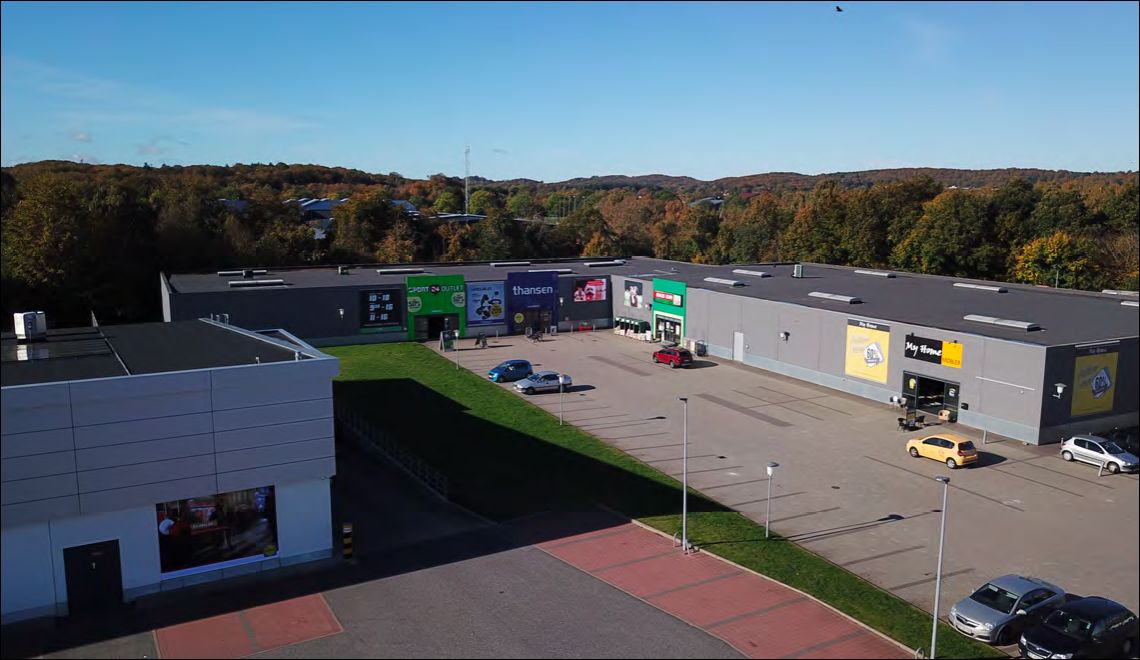Store bygninger med plakater og reklamer står på Retailpark Aabenraa. Parkplads med biler og spor efter parkering. Skove og
