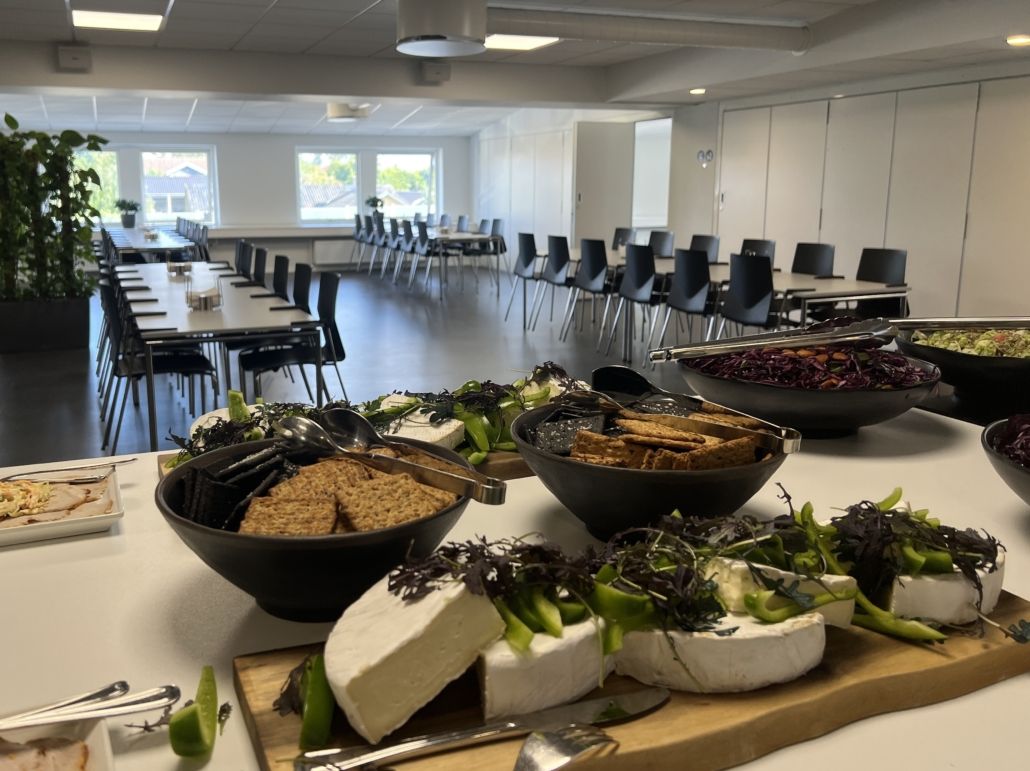 En stor møde- og konferencelokale med flere bord og stole samt en stor serveringsbord med diverse snacks og smørrebrød