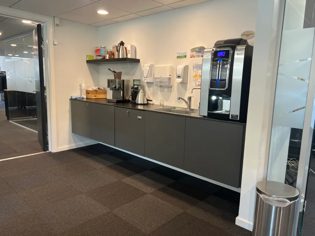 Et moderne kontor med en hvid væg og grå loft. Der er en kaffemaskine, en køkkenblandingsmaskine og en stålskraldspande. Der