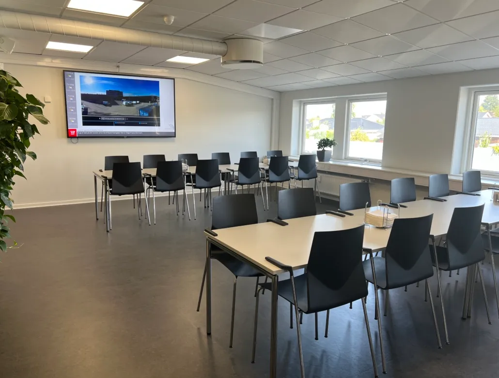 Et kontor med hvide vægge og gulv, store vinduer og et stort tv-skærm. Der er store runde borde med stole samt en stor grøn