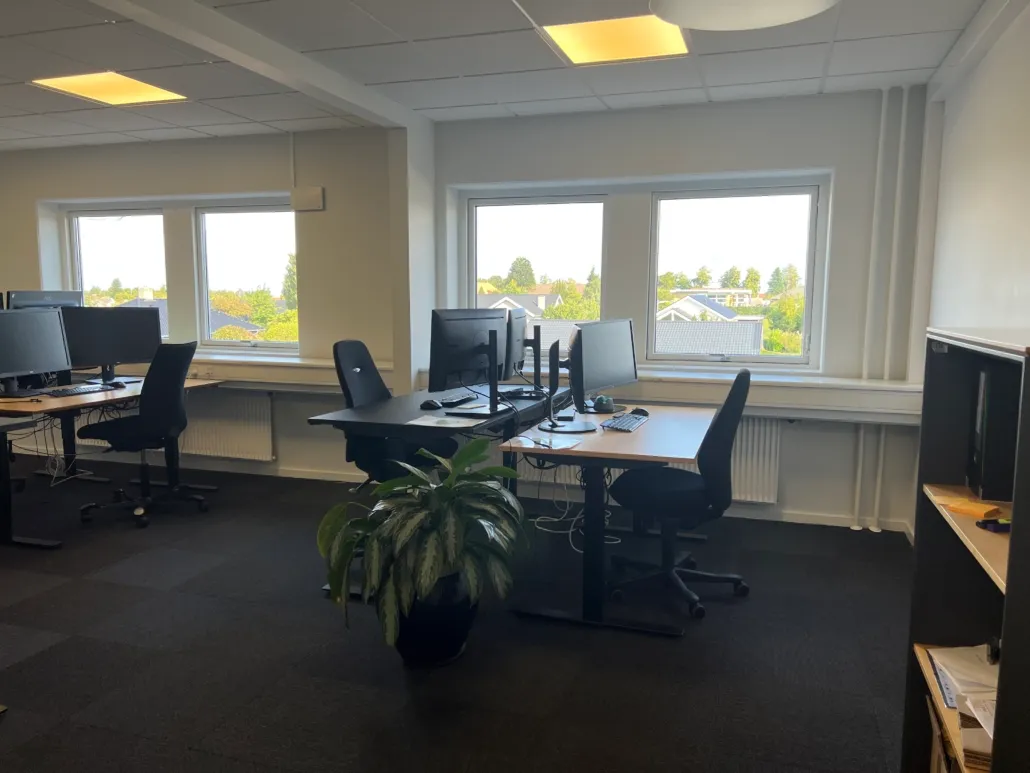 Et kontor med flere borde, hver med to computere, en plante og to lænestol. Der er tre vinduer med udsigt over et landskab.