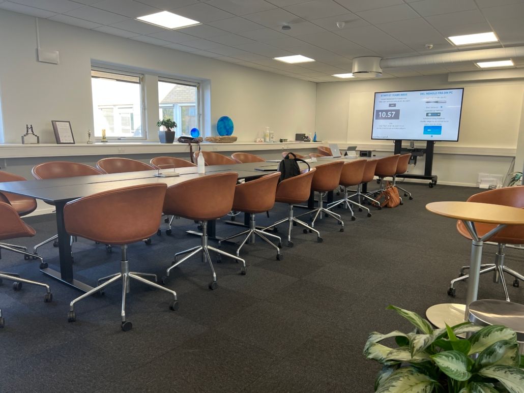 En konferencesal med fire rækker med læderstole, to lange borde og en stor tv-skærm i baggrunden