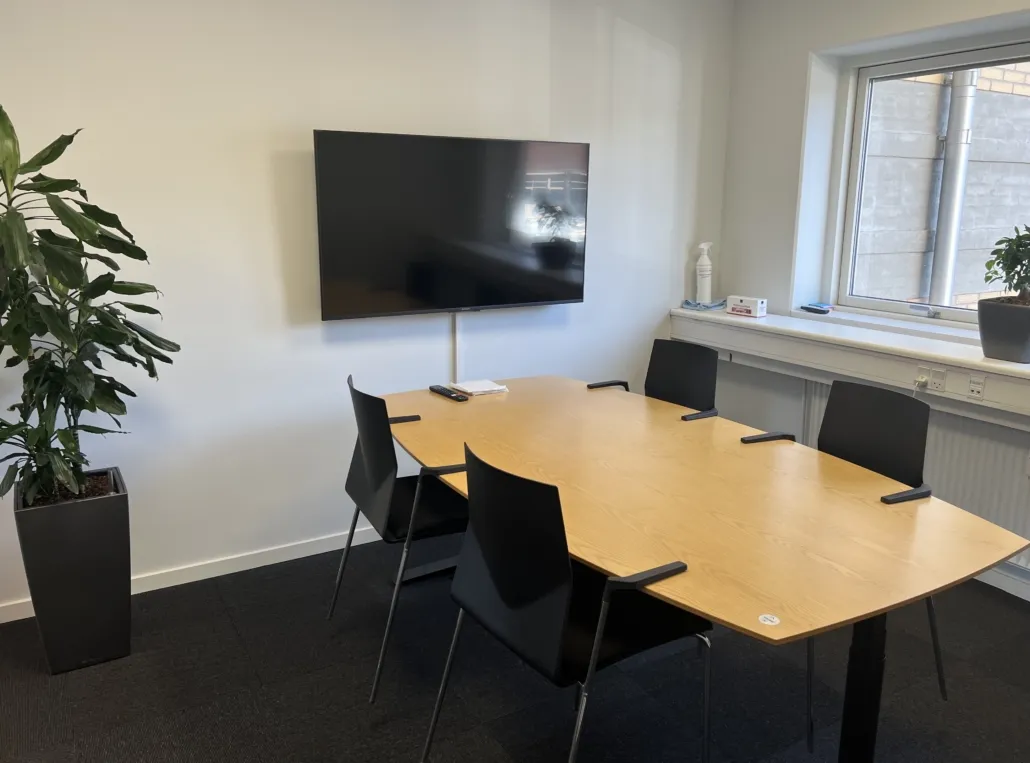 En konferencesal med gul bordplade, sort skærm og fire sorte stole, to store planter og et vindueskarmen med plante og