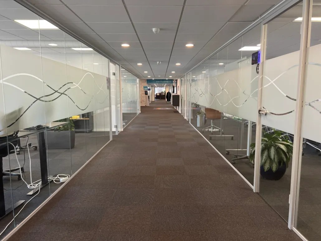 Kort og bred korridor med glasvægge og højloft. Væggene har skrift med svingende linjer og planter i gryder på gulvet