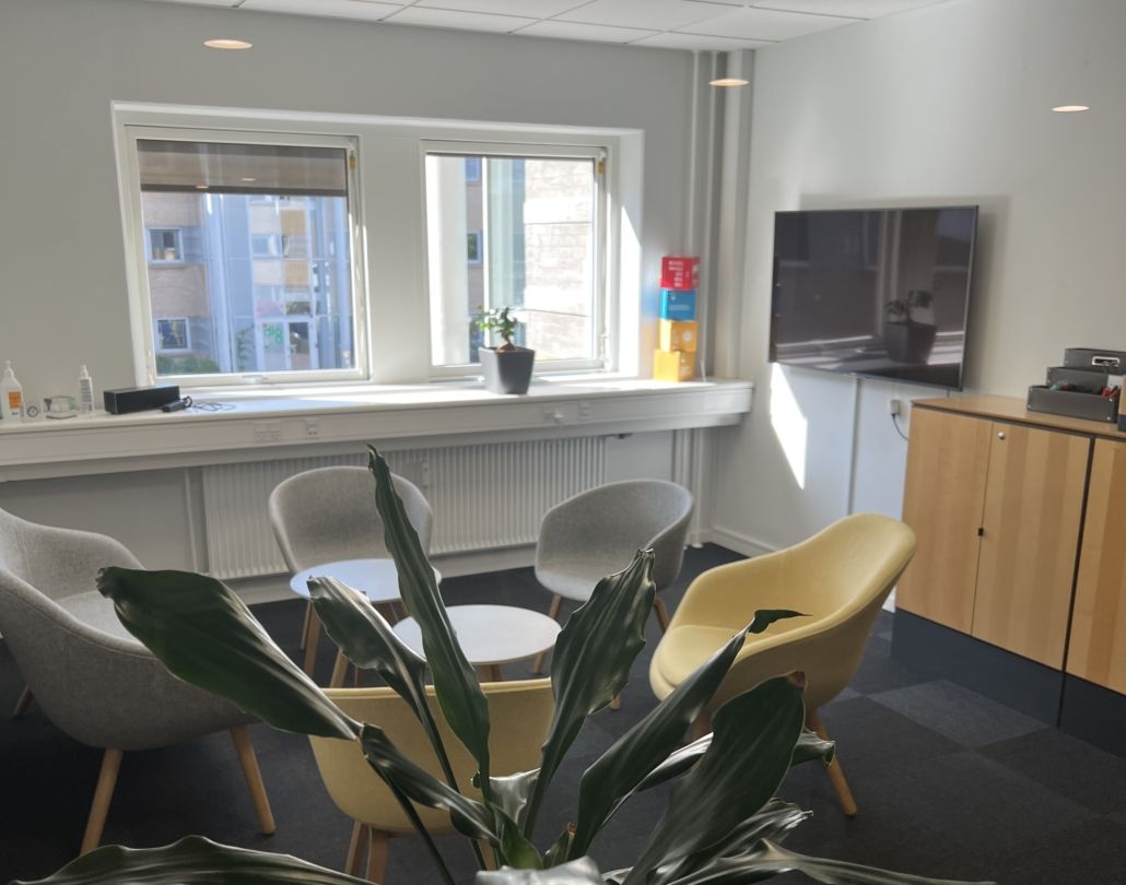 En kontor med hvide vægge, højloft og lyse gulve. Der er møbler og udstyr samt plante. Der er vinduer med udsigt til byen.