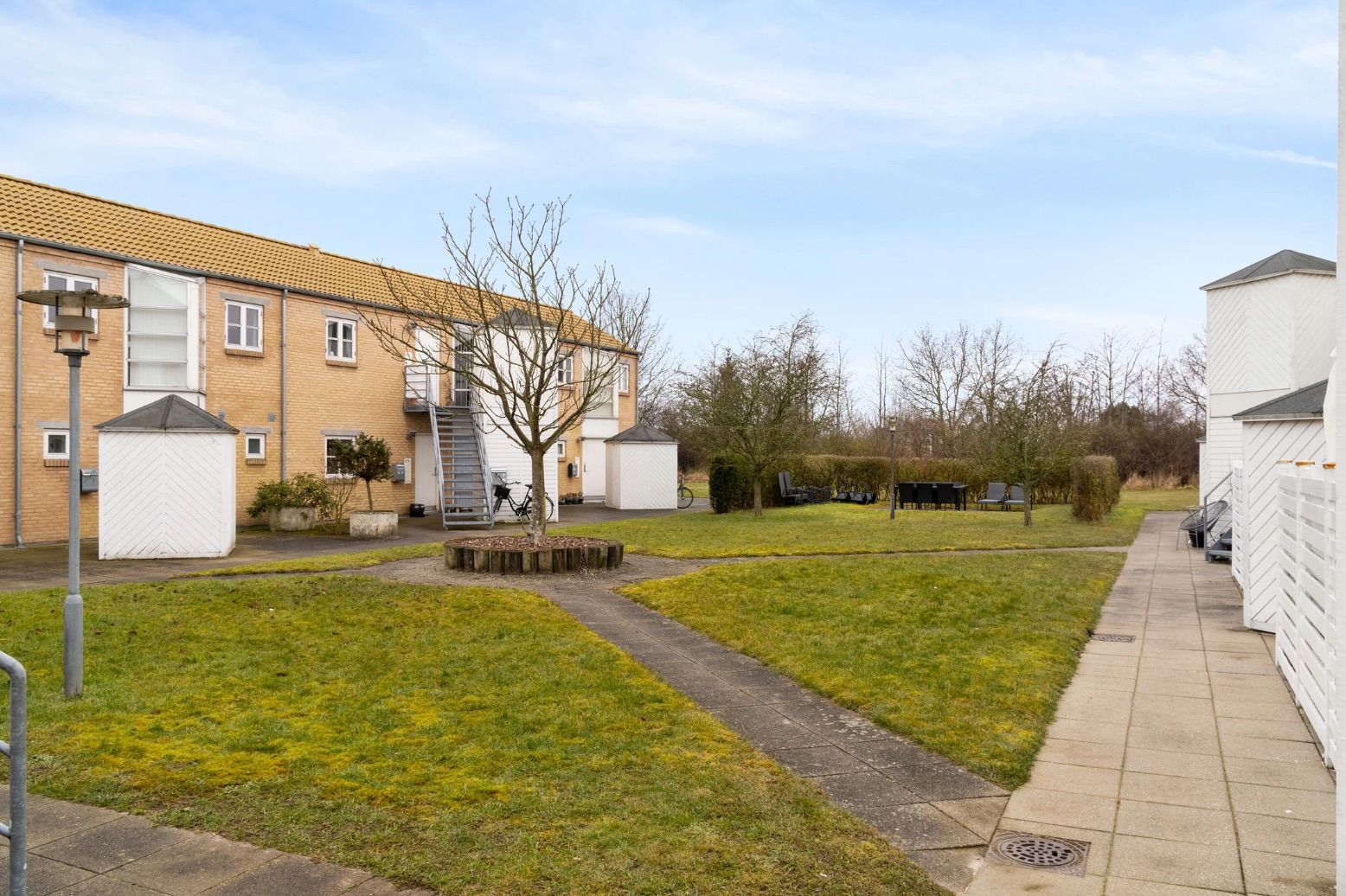 Lejlighedsbyggeri med fællesarealer og græsplæne, flere huse med terrasser og trapper, skur og møbelsæt med stole og bord,