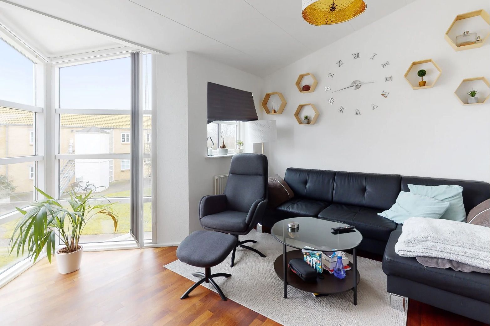 En lys stue med gult lys, en sort sofa med puder og en hvid tæppe på trægulvet