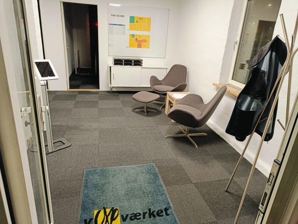 en ren kontorrum med en vægtekst “Find os her” og et skrivebord med en computer. Der er også en væghængende tøjstativ og en