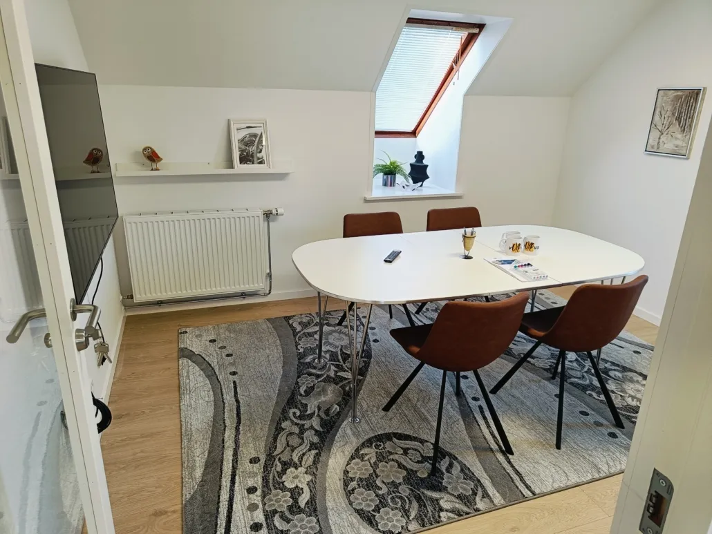 Moderne kontor med hvide vægge, lyse trægulve, rektangulært hvidt bord og fire brune stole med metalben. På bordet er der en