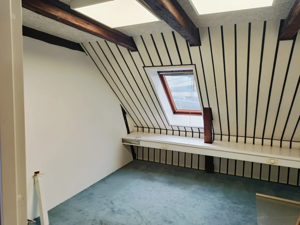 Værelse med loftsvinduer og loftsbjælke samt skrå gulv og hvide vægge