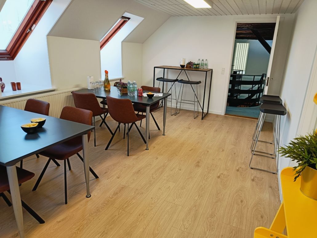 Værelse med højt til loftet, lyse loftsvinduer og moderne møbler som borde, stole og hylder. Bord med drikkevarer og snacks