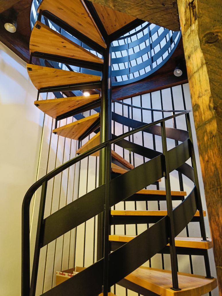 Spiralformet stige med lyse trætrin og mørke stålramme, placeret i et rum med hvide vægge og træbjælker