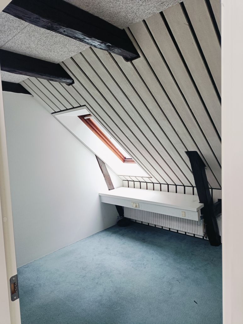 Loftsrum med højloft, lysegrå vægge, lyseblå gulv, skrånende loft og vinduesskarm i træ.