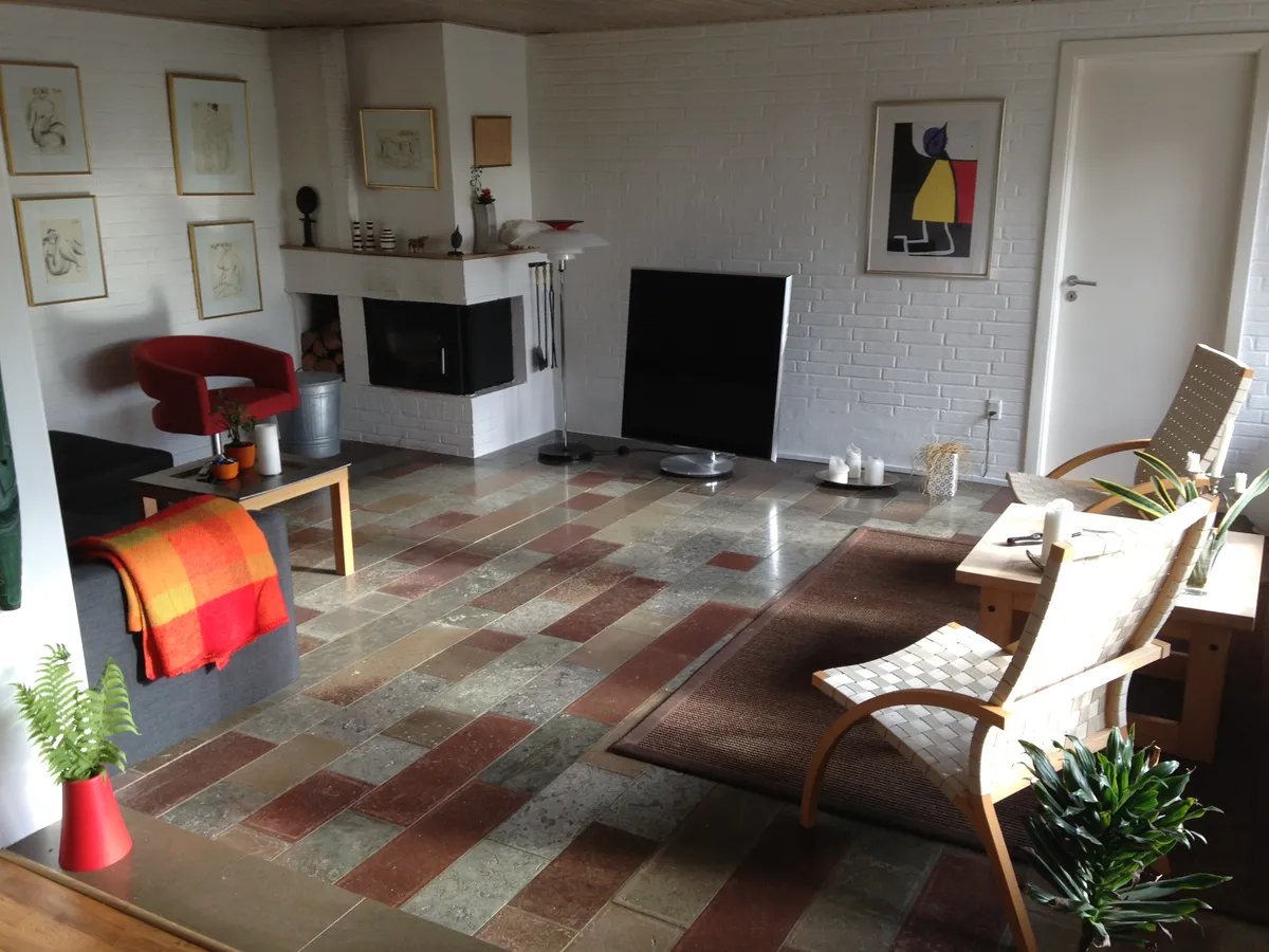 Stue med sofa, stole, trægulve, gulvtæppe, fjernsyn, hylde, malerier og køkken med pejs og loftsbjælker