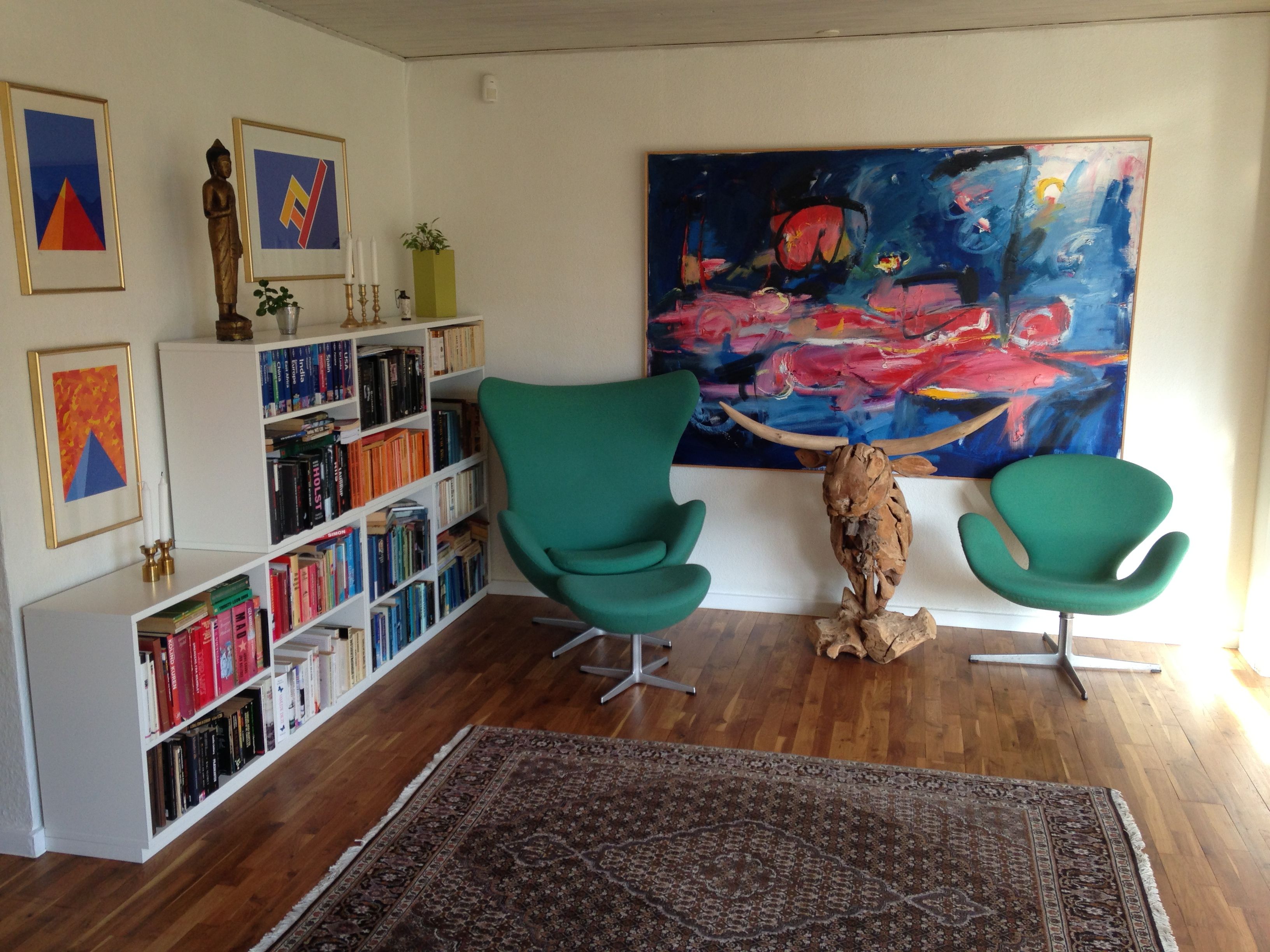 En hvid bogreol med mange bøger, to grønne lænestol og et stort maleri på væggen i et lyse og renslibet dansk interiør