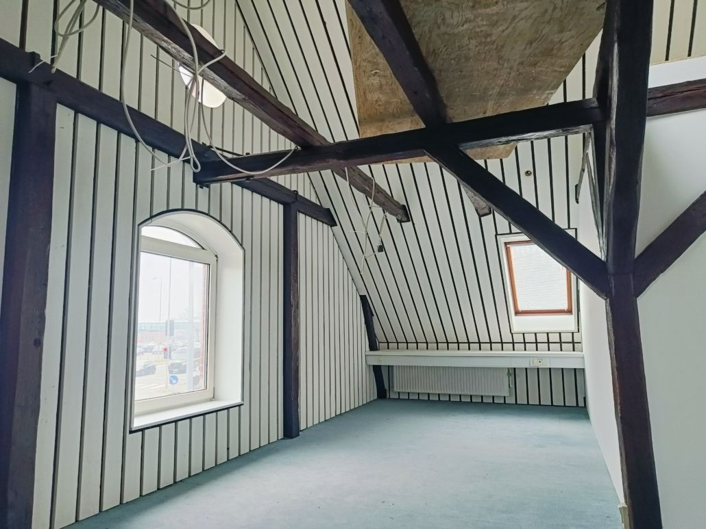 Foto af et loft med bærende bjælker og skrå stilk og vindue i væggen med udsigt over havnen