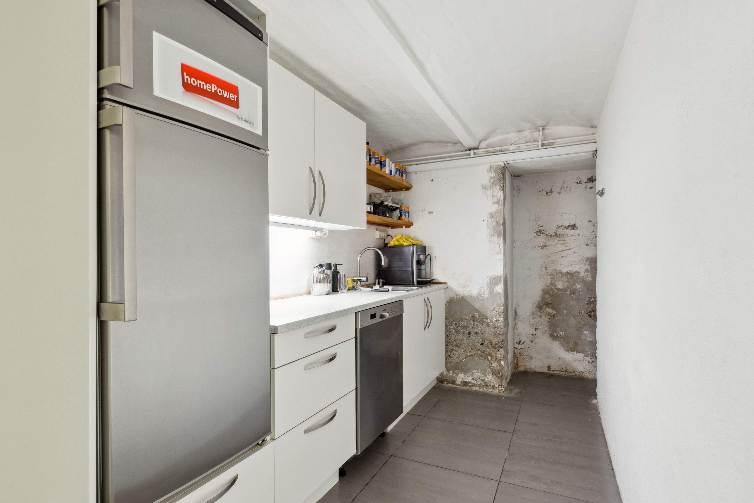 En lukket køleskab med HomePower-logoet og en rusten væg i et renoveringsbedyrvet køkken med et vaskemaskine, ovn og håndvask