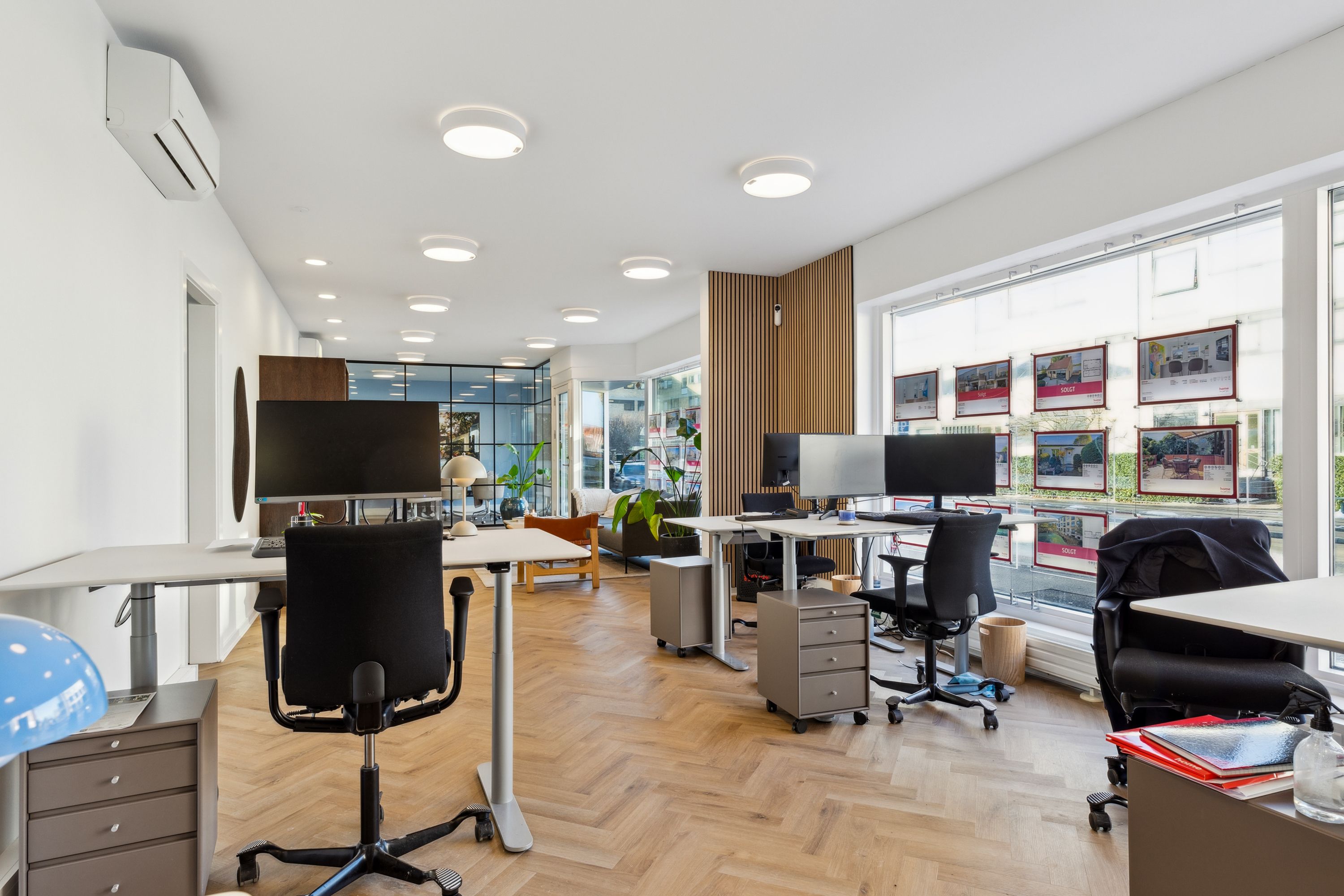 Kontorrum med høje vinduer, trægulve, lyse loftslamper og skriveborde med skærme og stole