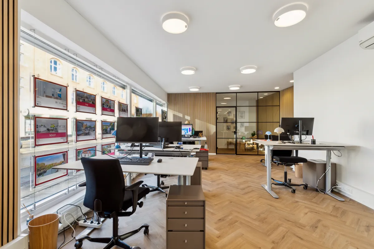 Kontor med skrivebord, stol, computer, luftkøler og udsigt til gaden med bygninger og skyer