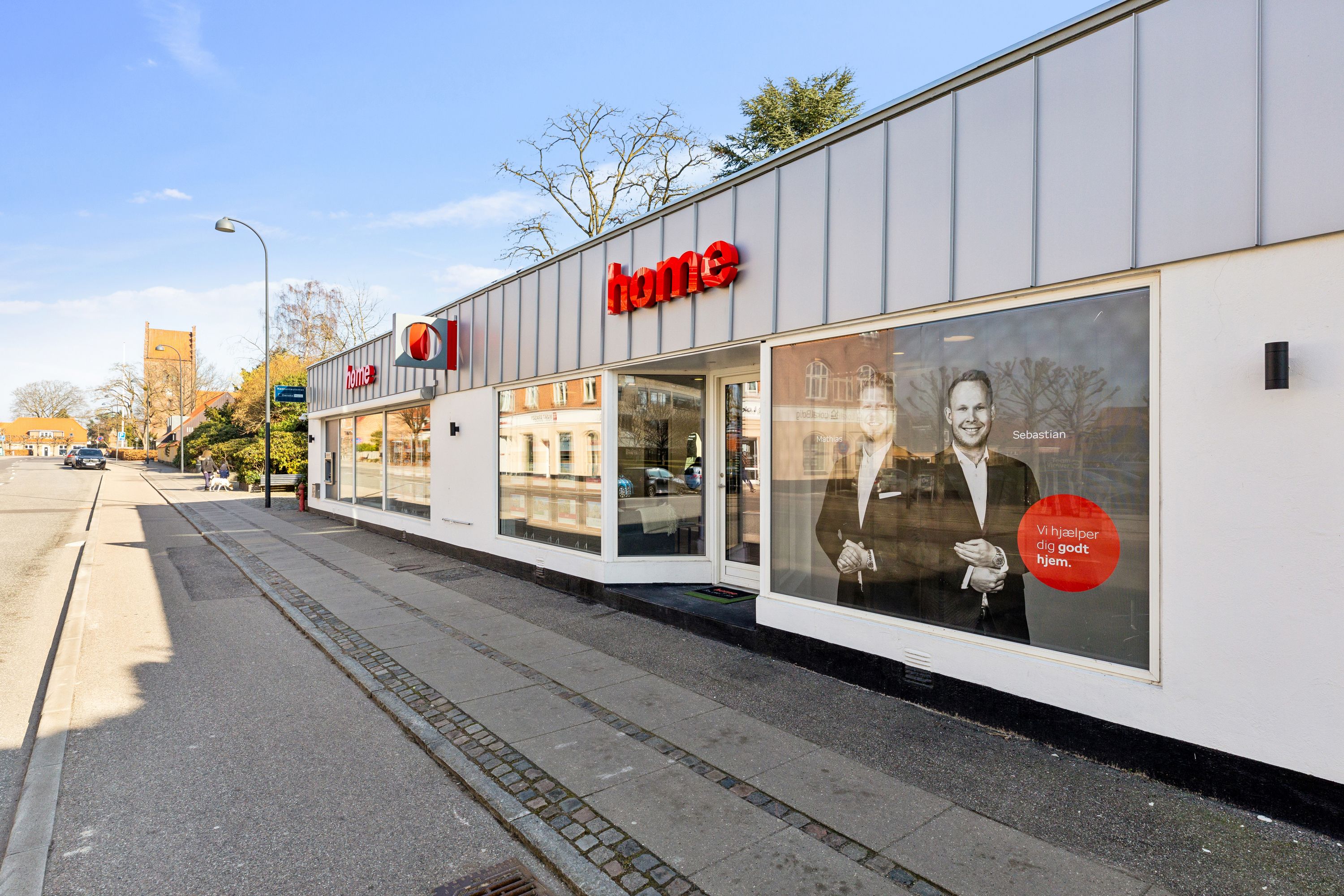 Butiksfront for Home A/S med stort vindue med reklameskilt og tekst "Vi hjælper dig godt hjem" samt portræt af to mand i