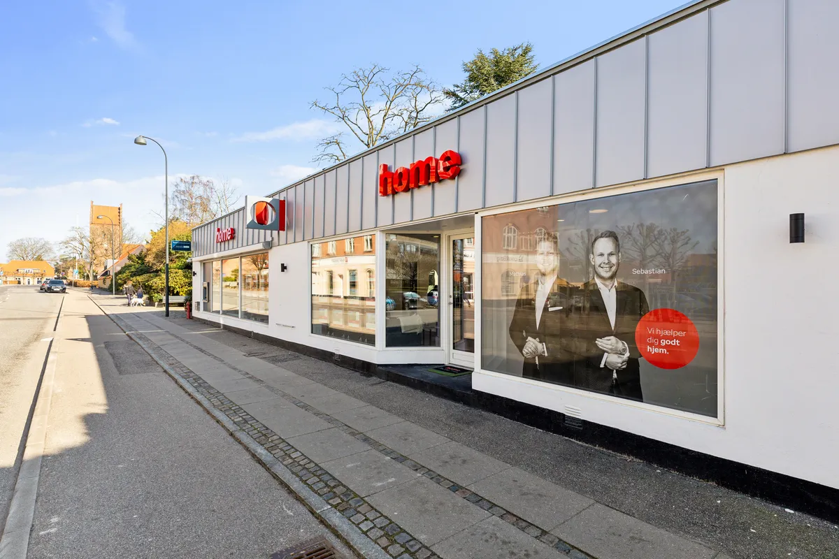 Butiksfront for Home A/S med stort vindue med reklameskilt og tekst "Vi hjælper dig godt hjem" samt portræt af to mand i