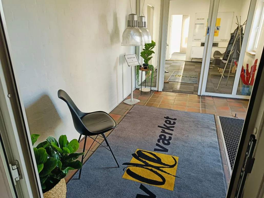 En hyggelig indgangszone med en lysegul gulvmåtte med logoet "Øe Værket" og en sort stol med plante og receptionsskilt