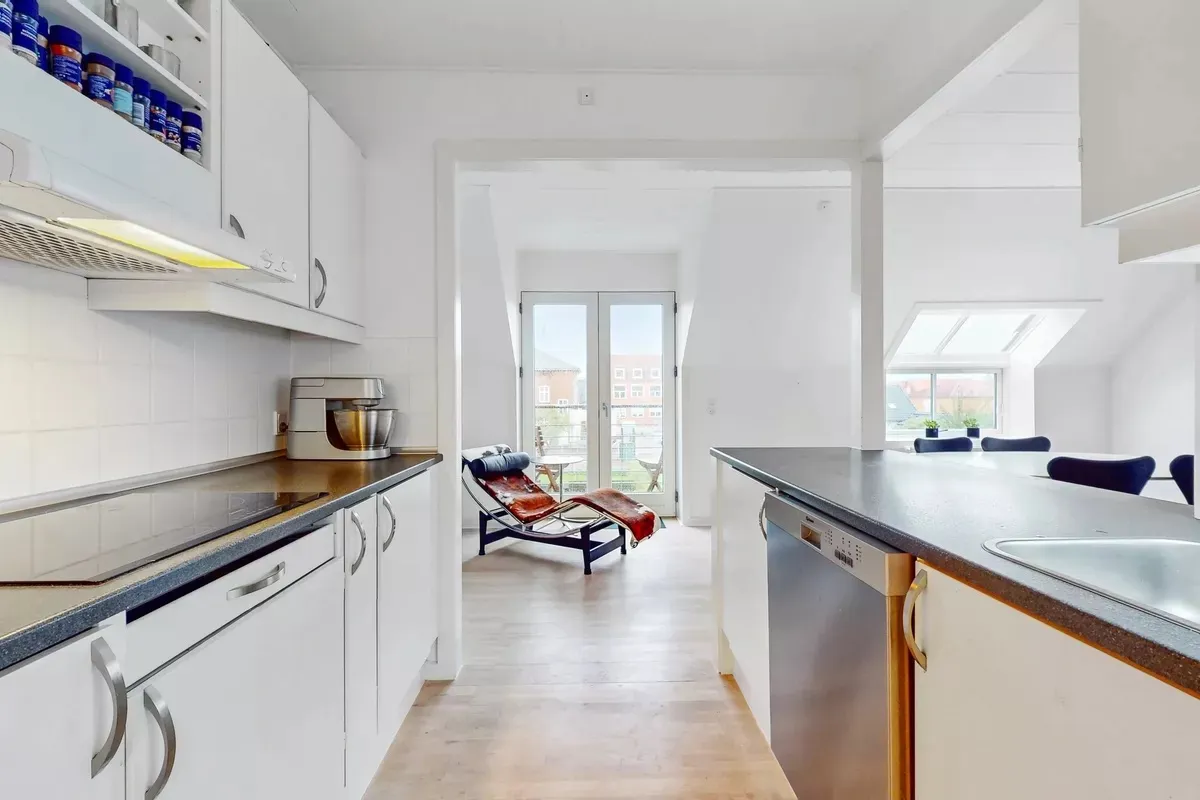 Moderne køkken med hvide vægge, trægulv, lysevinduer og møbler samt apparater som kaffemaskine og ovn