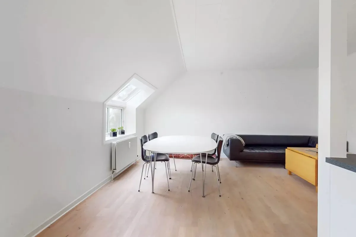 Indvendigt er der et rum med hvidt loft, højt vindue og en rund spisebord med fire stole samt en gul sofa med tæppe