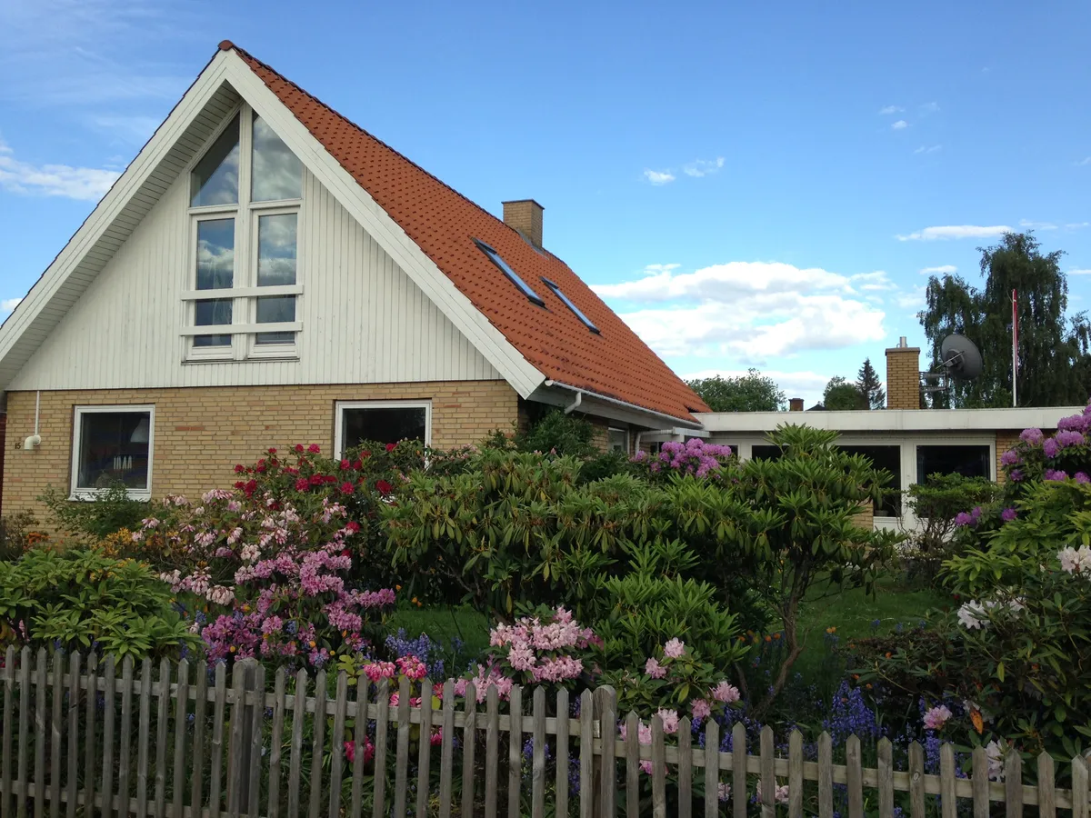 Villa med rødt tag og vinduer og have med røde og hvide blomster og grønne planter bag en hvid lav skur