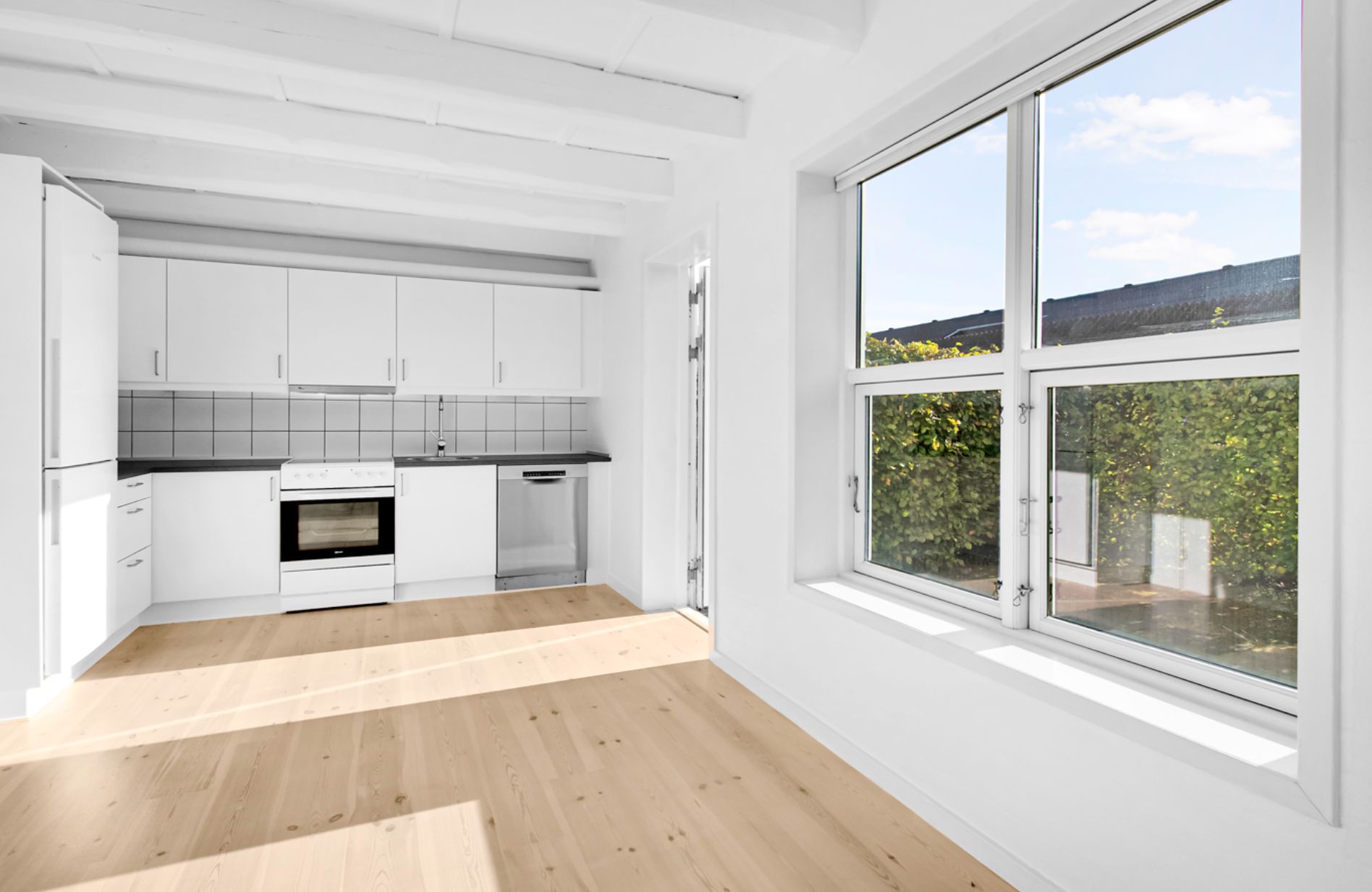 Lyst, moderne køkken med hvide vægge, hvidt køkkenskab, gulv og loft, indbygget ovn, komfur, spidskøkken og lyse vinduer med