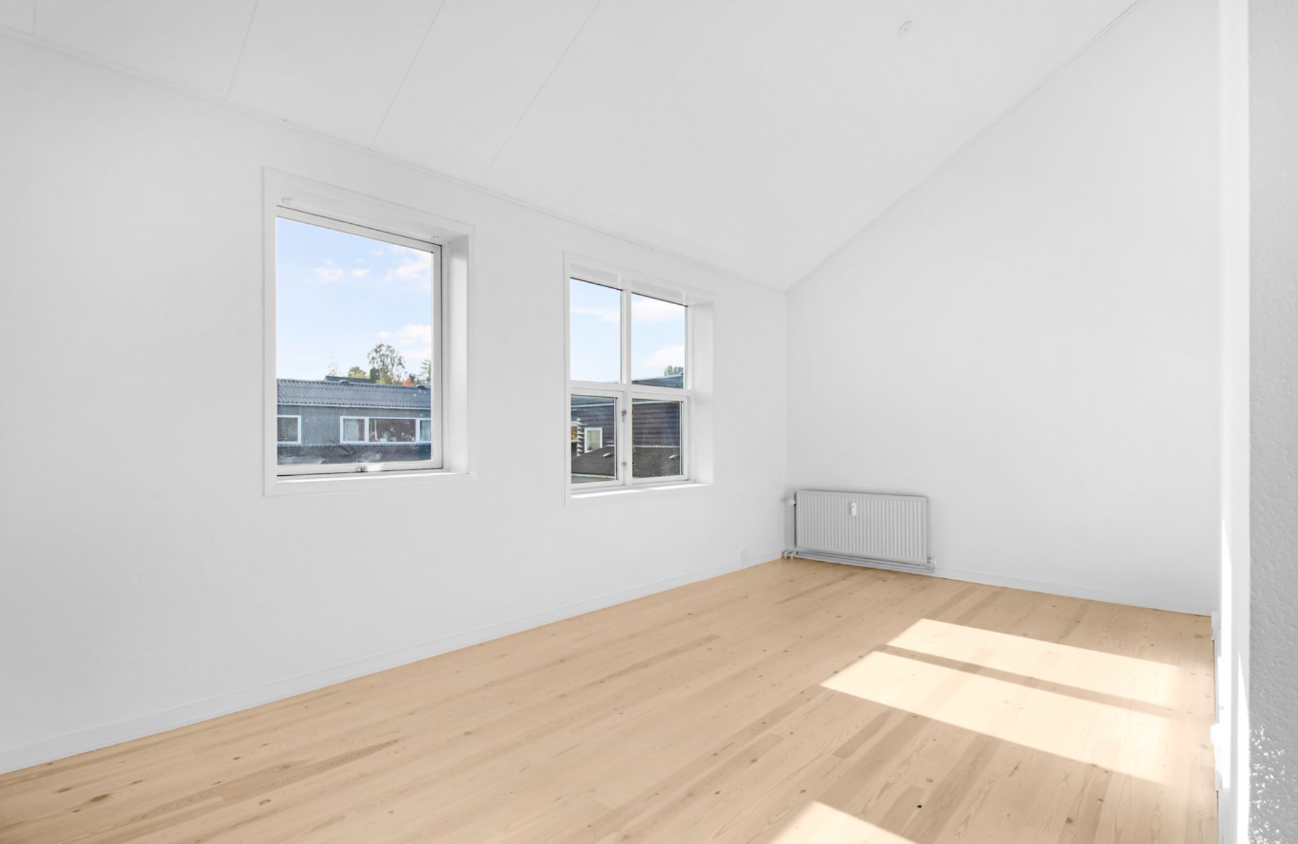 Lyst værelse med lyse, trægulve og to store ruder mod syd. Væggene er hvide, og der er en hvid radiator på den ene væg.