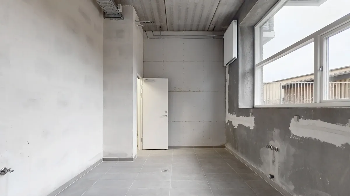 En stor, tom og grå bygning med to døre og en stor vinduesåbning. Gulvet er glat og bleggråt og væggene er hvide og grå.