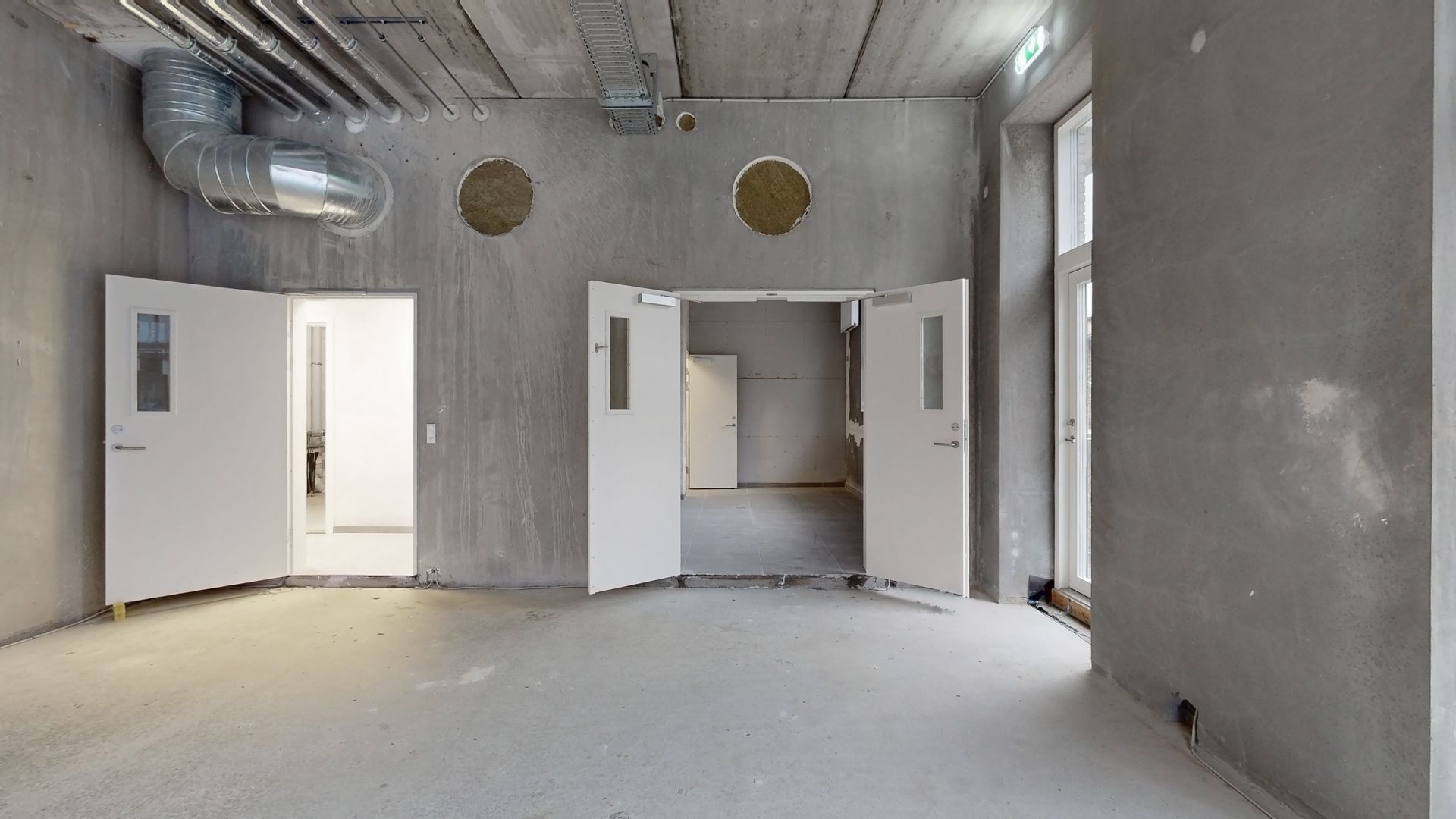 En tom rum med tre åbne hvide døre, en stor rørledning i loftet og lysegrå vægge med to runde åbninger