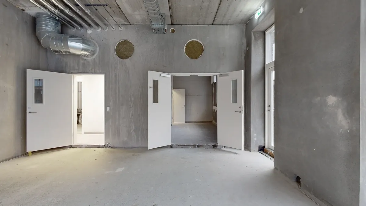 En tom rum med tre åbne hvide døre, en stor rørledning i loftet og lysegrå vægge med to runde åbninger