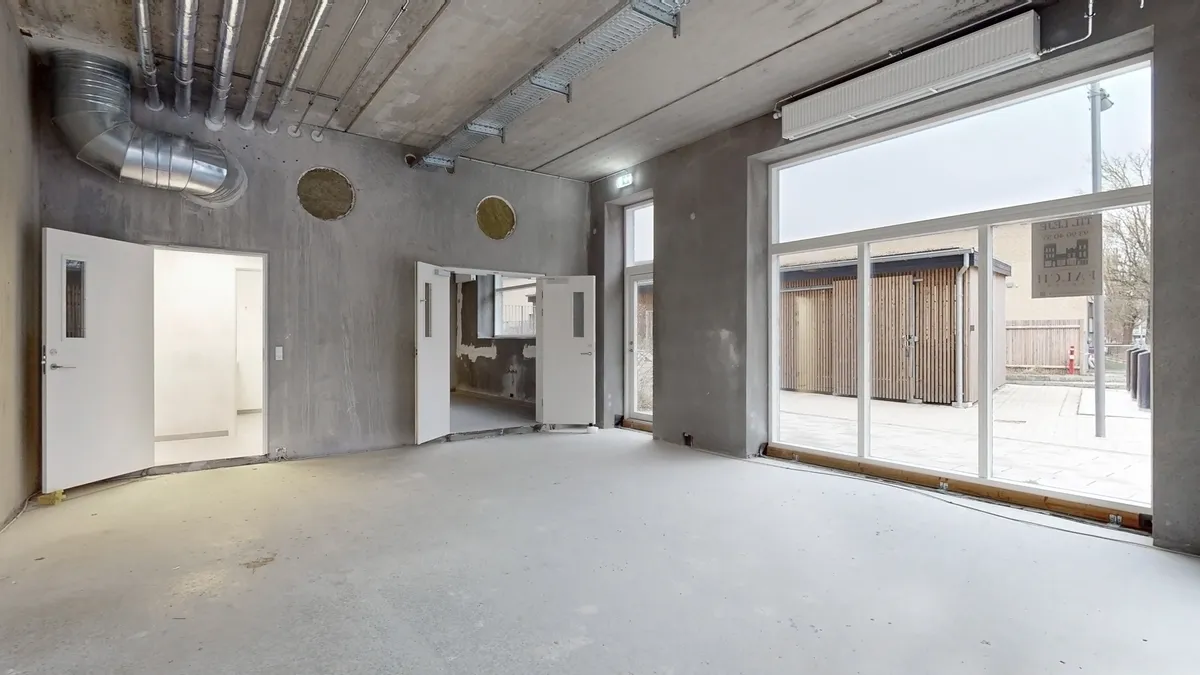 Et stort lokale med åbne døre og store vinduer til en gård. Pladsen er tom og der er en væg med åbninger og rør. Pladsen har