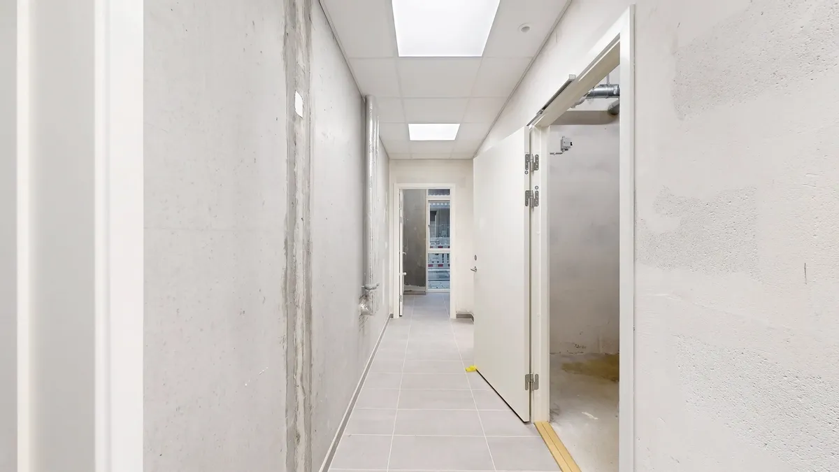 Et langt og smalt, lysegråt og pænt hulrum med hvide vægge, gulv og loft samt åbne døre og en indgangsdør i hvidt og sølv