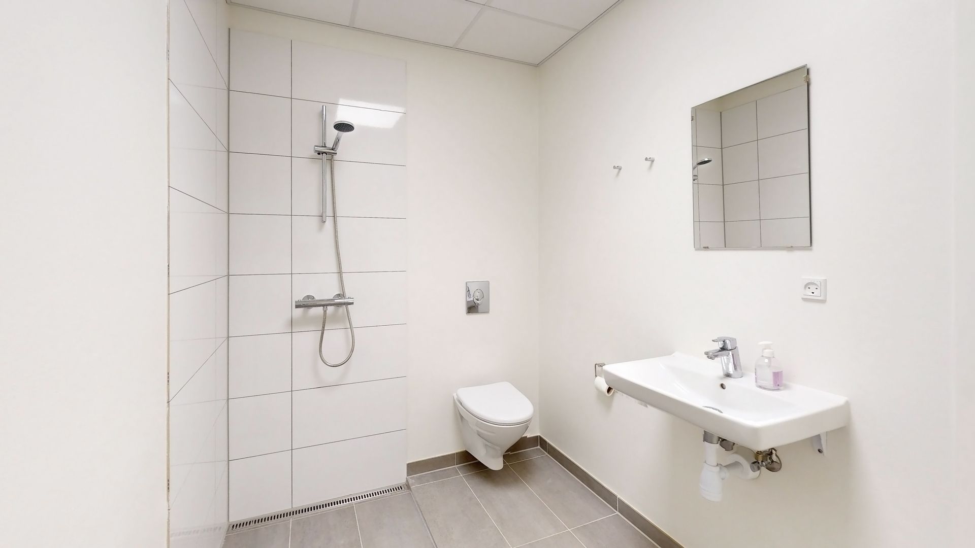Et moderne, hvidt badeværelse med højloft og hvidt toilet og håndvask med spejl samt brusevæg med hængende brusehoved