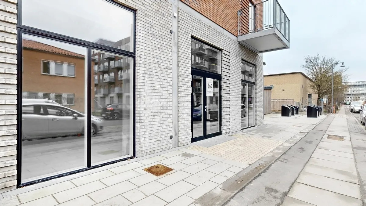 Glassdøre med skilte på facaden af en ny bygning med en balkon og parkerede biler og søjler langs gaden