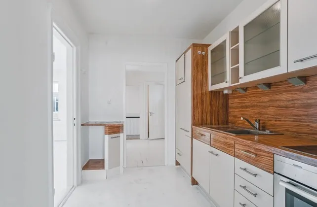 Minimalistisk køkken med hvide vægge og lysegul gulv. Store skabe med hvide facader og træplade på væggen. Rustfrit stål