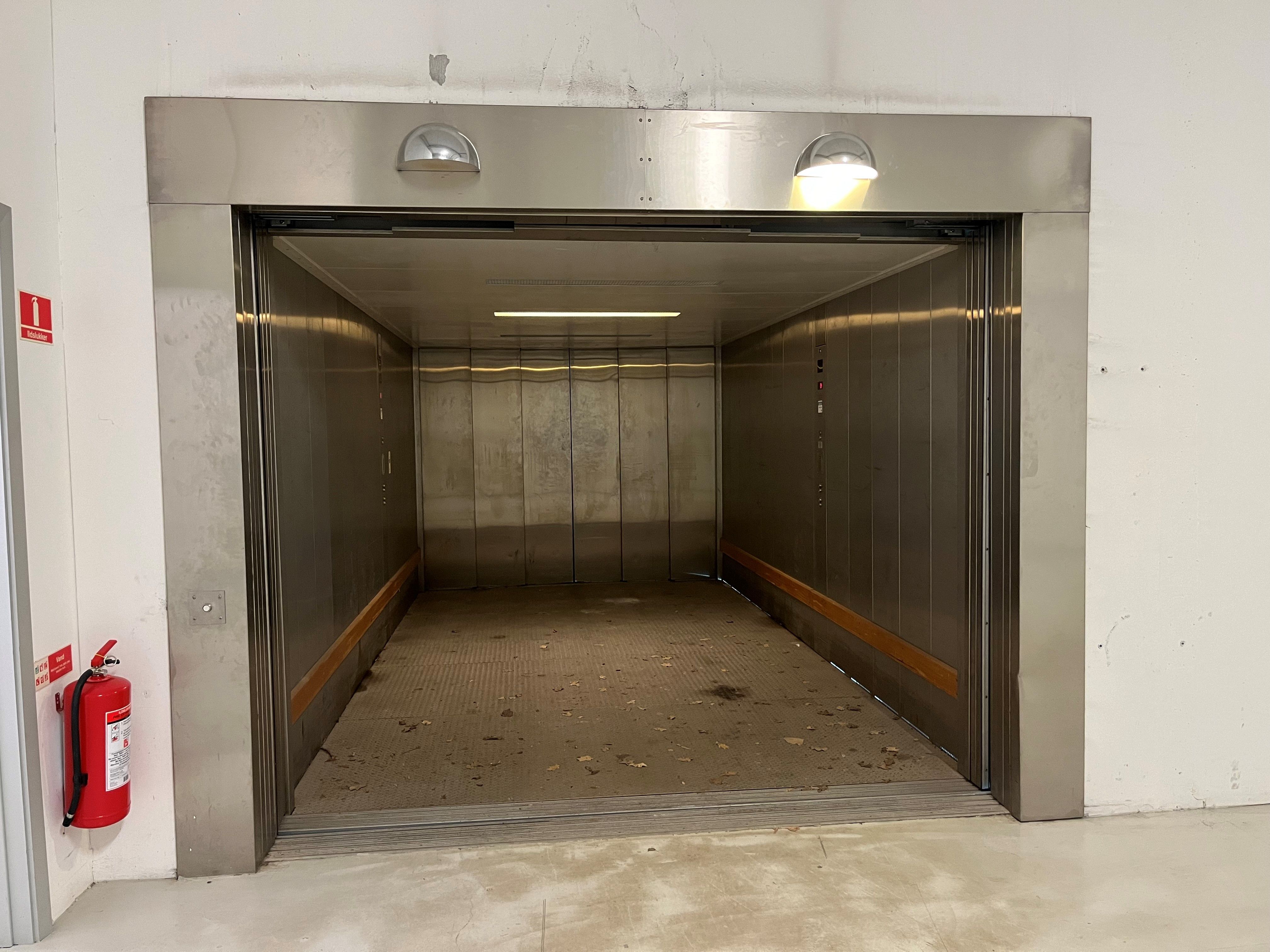 En stor vareelevator med åbne døre og et rødt brandslukker i baggrunden og høje lofter med lyse armaturer i taket