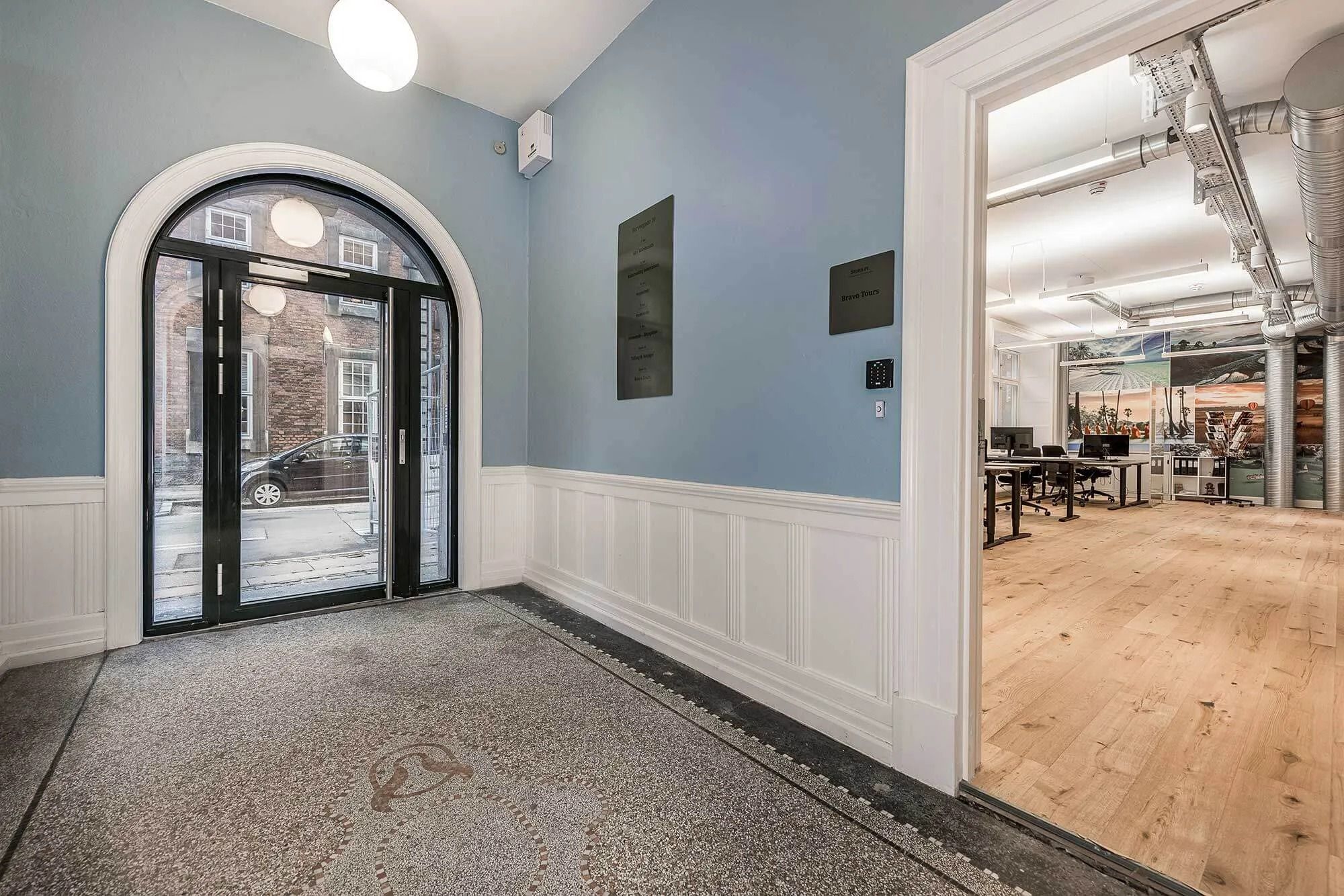 En nyrenoveret kontor med højloft, lyse gulve og moderne møbler, samt en stor, bueformet glasdør med udsigt til gaden