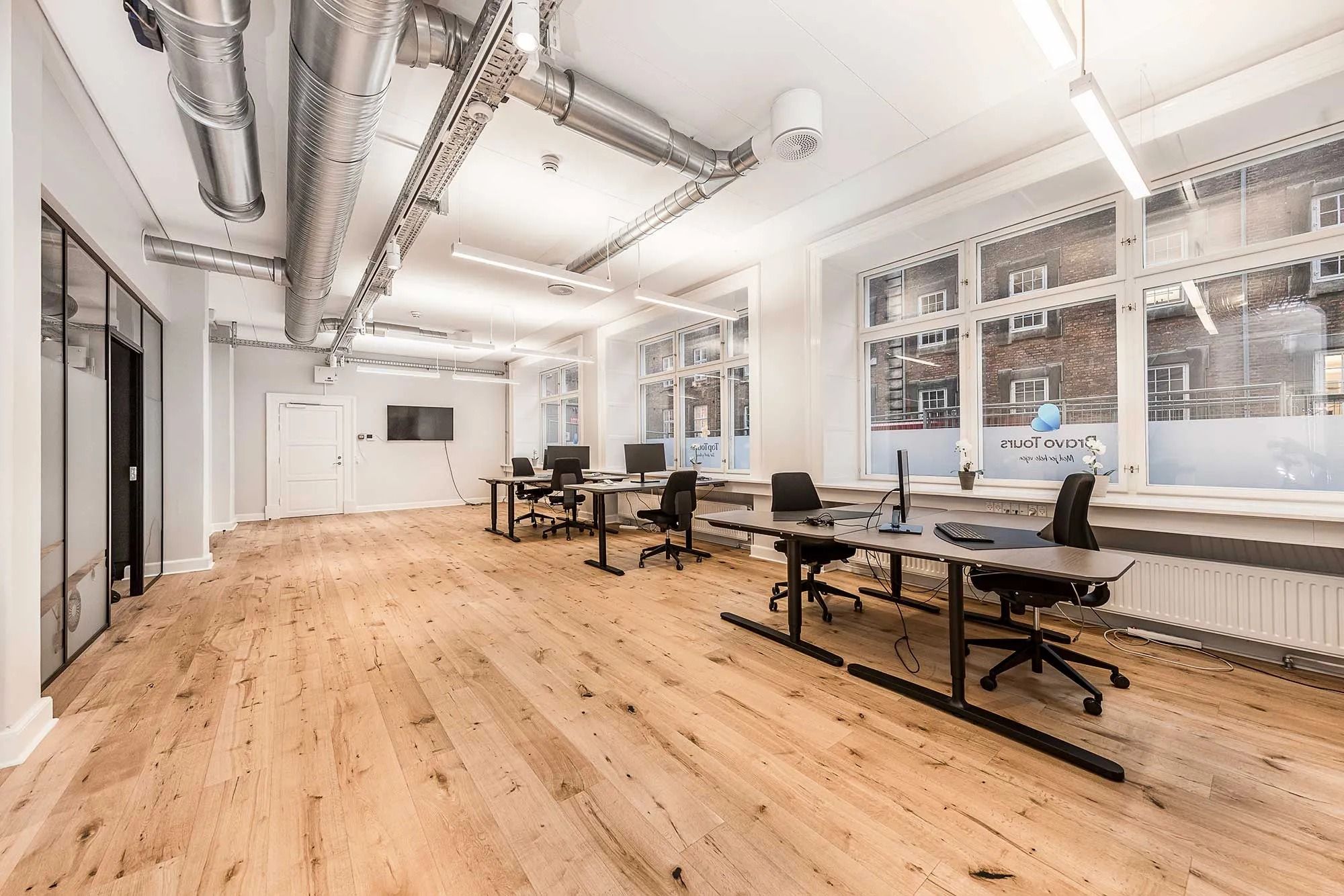 Kontor med lyse, høje vinduer, moderne møbler og hårde trægulve samt hvide vægge og store vinduer mod gaden