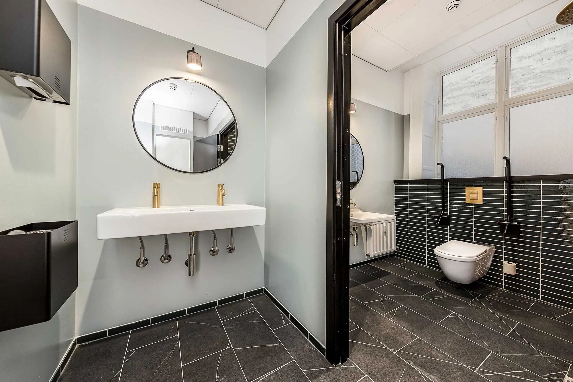 Moderne badeværelse med hvidt vasketøj, guldbeslåede vandhane og to toiletter, samt lysegrå vægge og sort marmorgulv