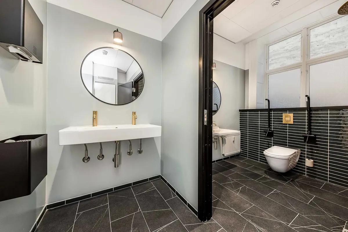 Moderne badeværelse med hvidt vasketøj, guldbeslåede vandhane og to toiletter, samt lysegrå vægge og sort marmorgulv