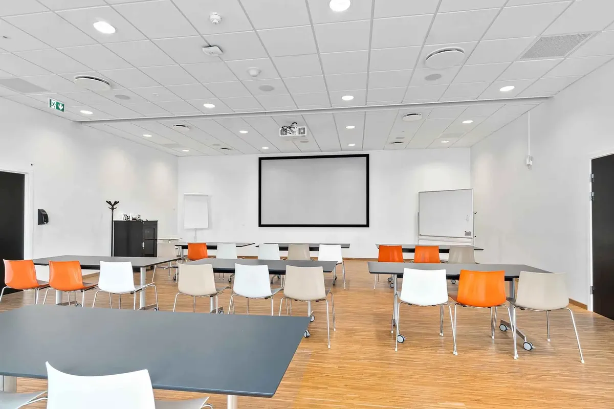 Moderne konferencelokale med hvide vægge og trægulv, hvor der er flere borde og stole, og et stort hvidt whiteboard og en