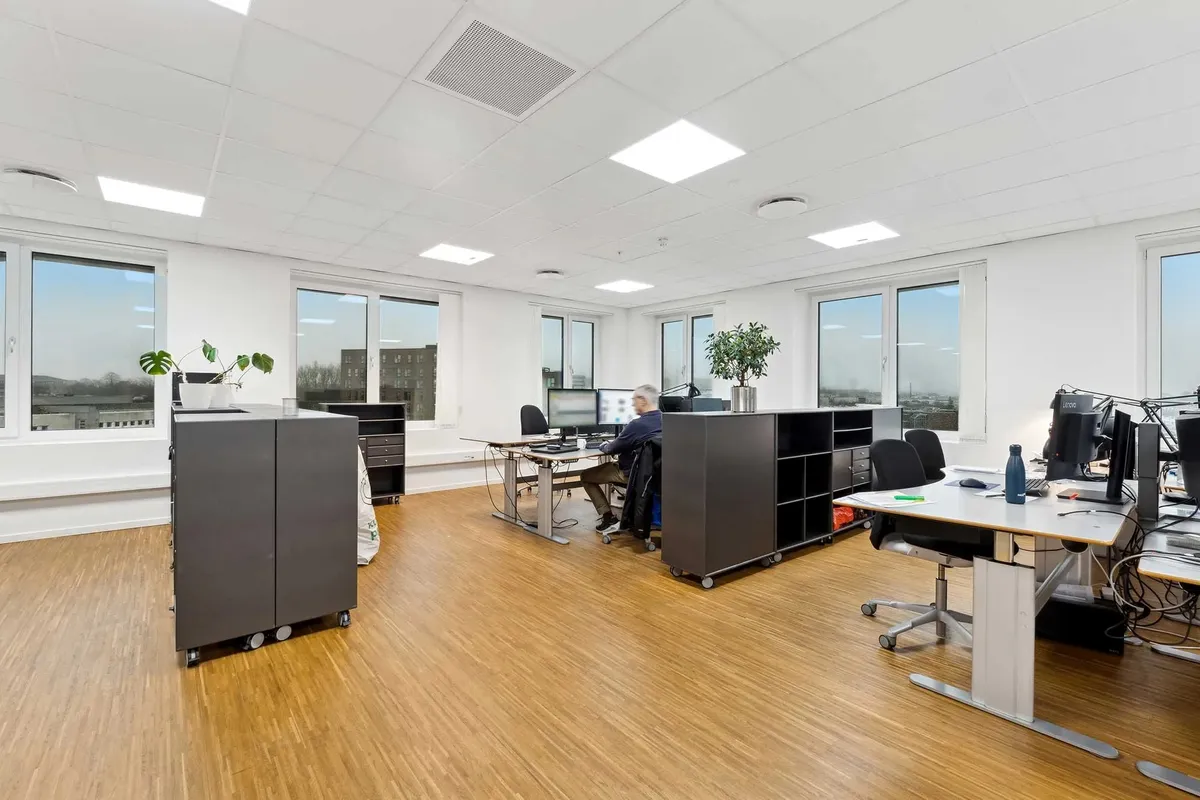 Et moderne kontor med lyse lofter og store vinduer med udsigt til byen. Der er flere arbejdsborde med computere og