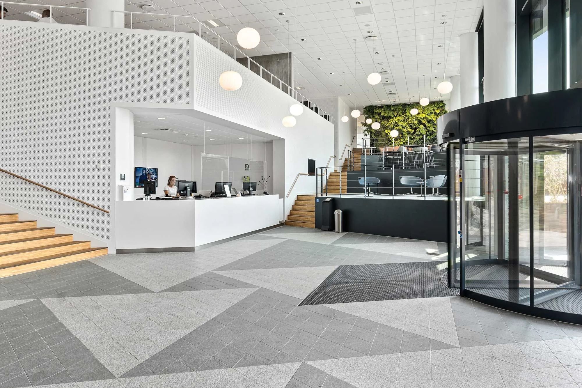 Moderne lobby med trapper, skrivebord og stol, glasrevolverdør og lysekroner, og en person sidder ved skrivebordet