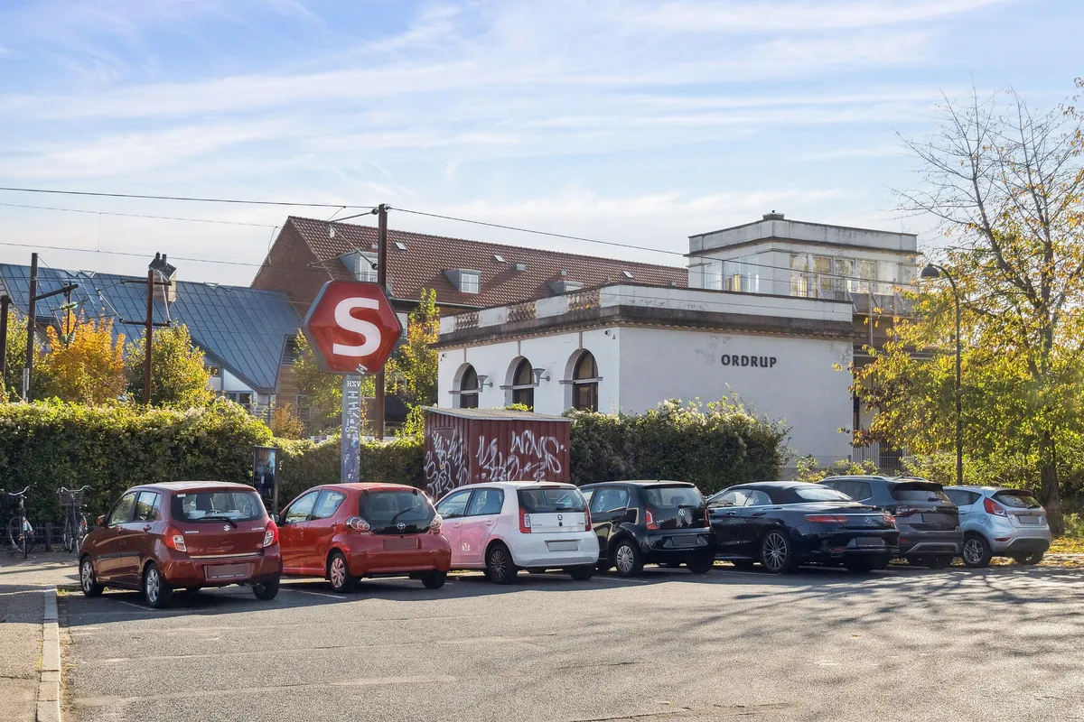 Parkerede biler i Charlottenlund med et stopskilt og Ordrup bygning i baggrunden. Foran er et stopskilt, og bagved er en bygning med teksten Ordrup.