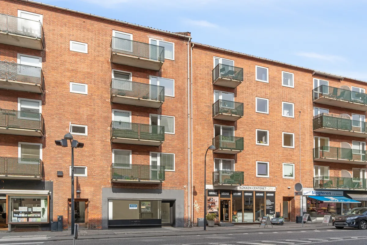 Butikslomme på Ordrupvej 64 med lagerrum og praktisk baglo. Lejlighedsbygning med balkoner og grønne områder. Parkeret bil og salgsskilt udenfor Hacker og Køkken Centert.
