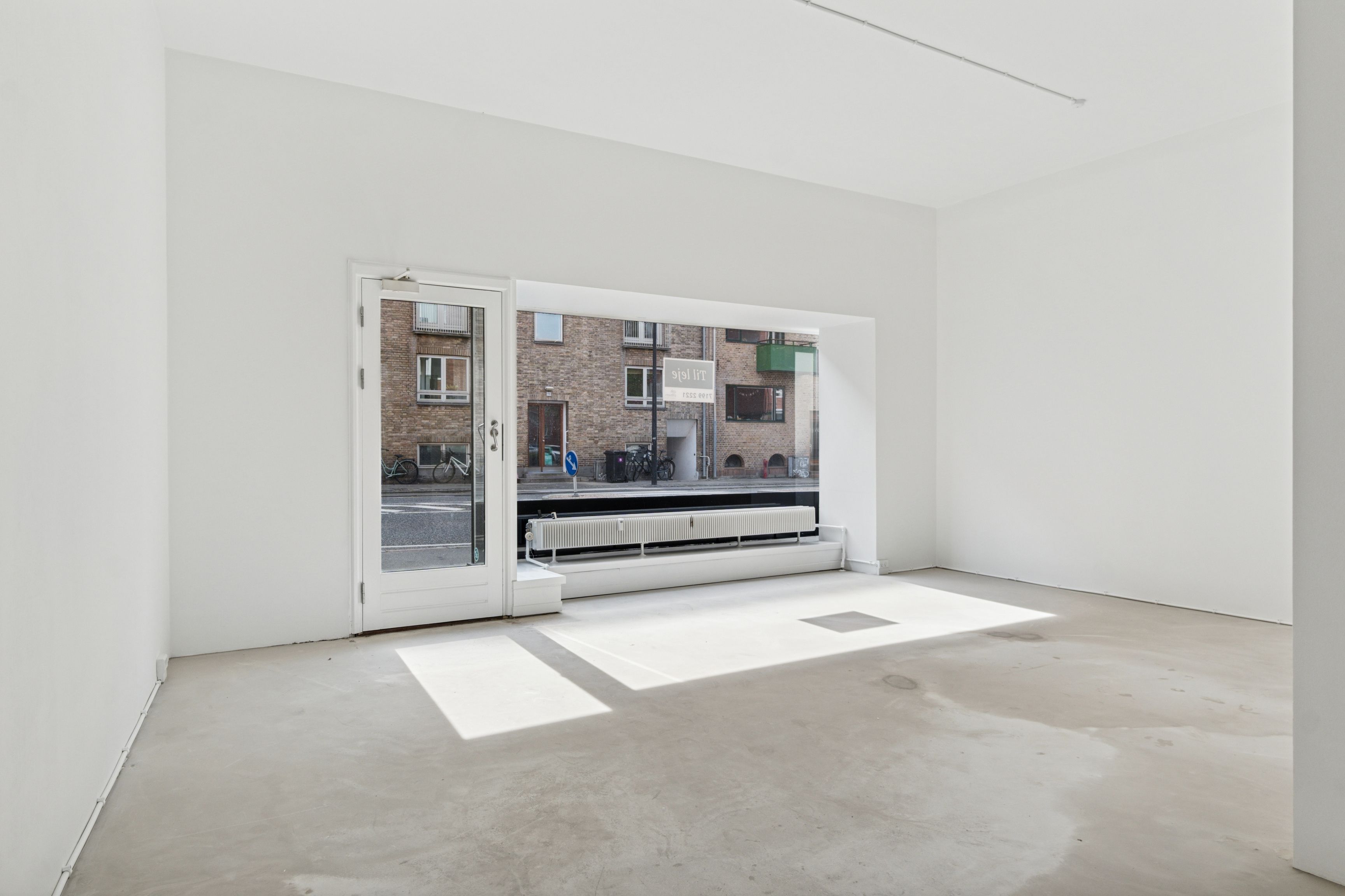 Lyst og rent butikslokale med hvide vægge og gulv og et stort glasvindu med udsigt til gaden med cykler og bygninger